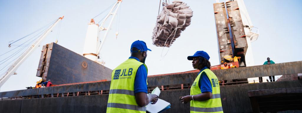 Deux agents de port en gilets jaunes discutent devant un navire de transport déchargeant des sacs de marchandises.