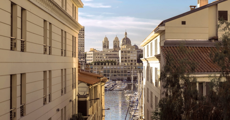 Vue d'un port de plaisance avec des bateaux, encadré par des bâtiments urbains et une cathédrale au loin.