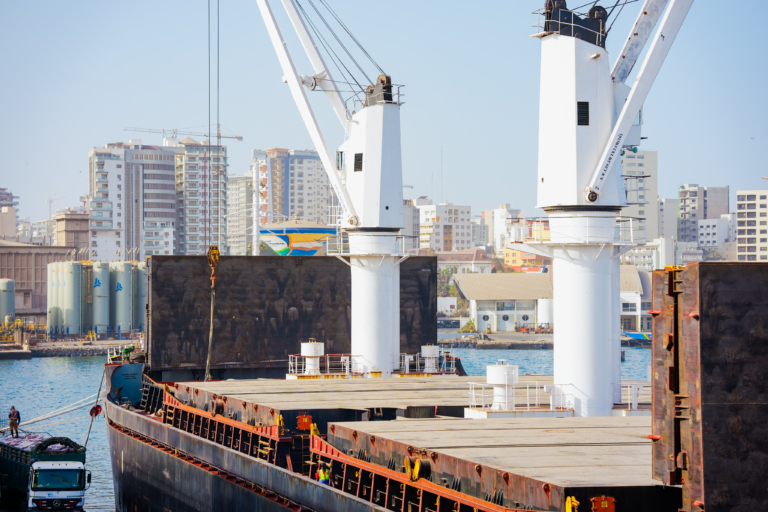 Grues blanches imposantes sur un cargo à quai dans un port avec des immeubles en arrière-plan.
