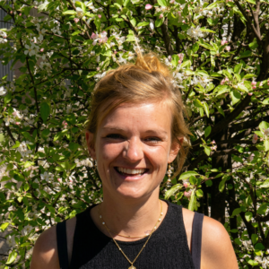 Une femme souriante aux cheveux blonds attachés pose devant un arbre en fleurs blanches et roses.