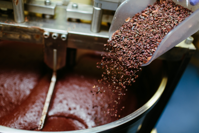 Des éclats de fèves de cacao sont versés d'une pelle dans un mélangeur industriel contenant du chocolat fondu.
