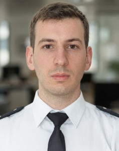 Portrait d'un homme en uniforme blanc avec une cravate noire, regardant fixement l'objectif.