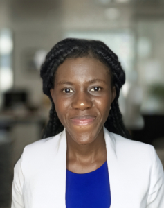 Portrait d'une femme noire souriante avec des tresses, portant un blazer blanc sur un haut bleu.