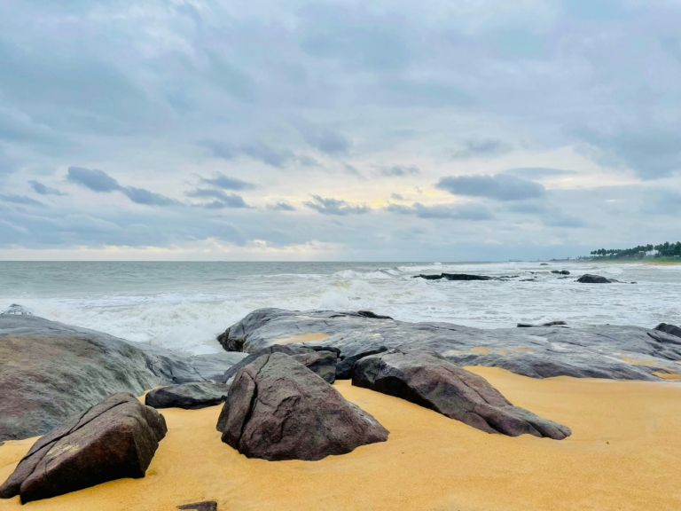 Vue d'une plage de sable doré avec des rochers sombres et des vagues blanches sous un ciel nuageux.