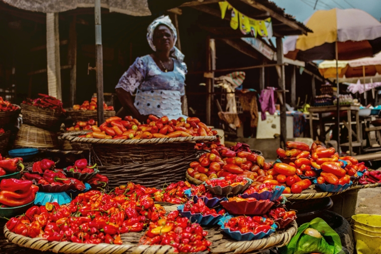 Une femme se tient derrière des paniers remplis de piments rouges et oranges sur un marché.