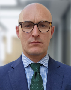 Portrait d'un homme chauve portant des lunettes, un costume bleu, une chemise à carreaux et une cravate verte.