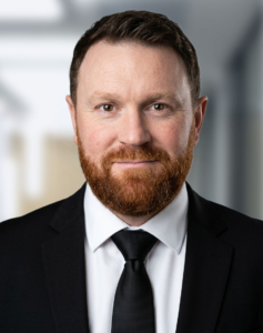 Portrait d'un homme à la barbe rousse portant un costume noir, une chemise blanche et une cravate noire.