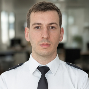 Portrait d'un homme en uniforme blanc avec une cravate noire, regardant fixement l'objectif.