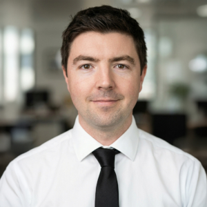 Portrait d'un homme souriant portant une chemise blanche et une cravate noire sur un fond de bureau flou.