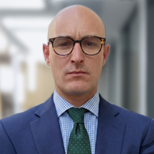 Portrait d'un homme chauve portant des lunettes, un costume bleu, une chemise à carreaux et une cravate verte.
