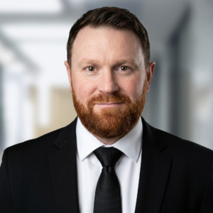 Portrait d'un homme avec une barbe rousse, portant un costume noir, une chemise blanche et une cravate noire.