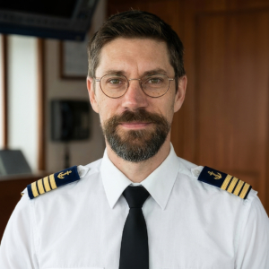 Portrait d'un homme avec des lunettes et une barbe, portant un uniforme blanc de capitaine de navire.